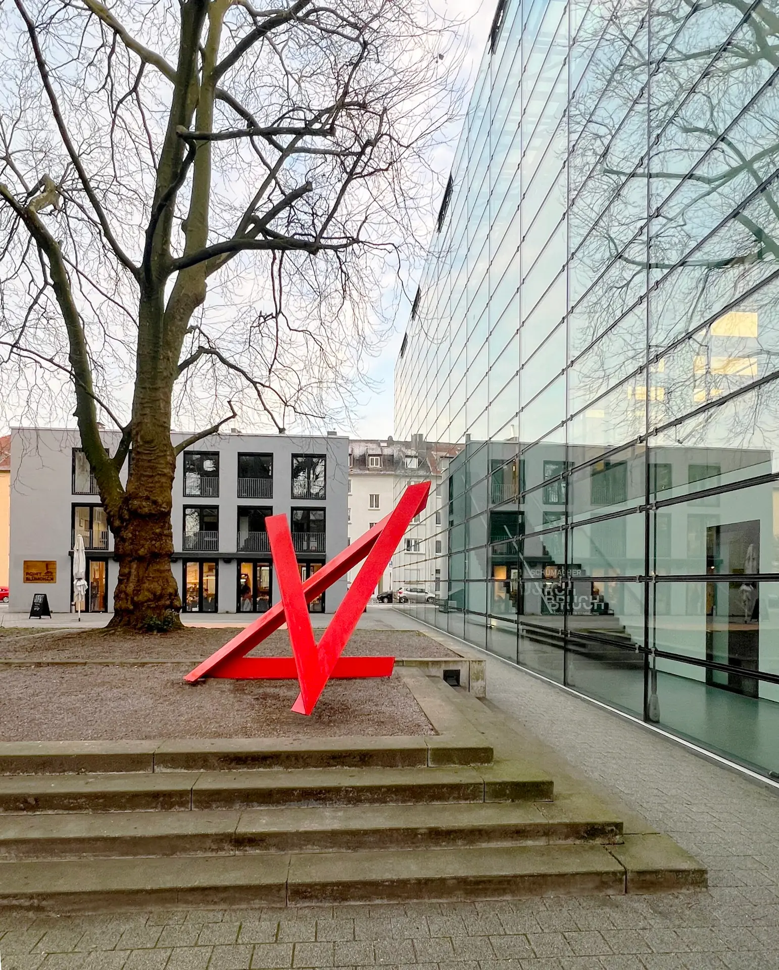 Rote abstrakte Skulptur aus Stahl, der linienhaft in verschiedene Richtungen zeigt von Utz Brocksieper vor der modernen Glasfassade des Emil Schumacher Museums neben einer großen Baumgruppe ohne Blätter