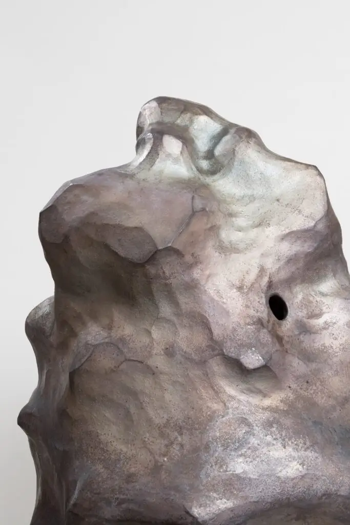 Die Skulptur hat organische Formen und erdige sowie metallische Farben. Sie läuft nach oben hin spitzer zu. Im oberen Teil befindet sich ein Loch. Detailansicht.